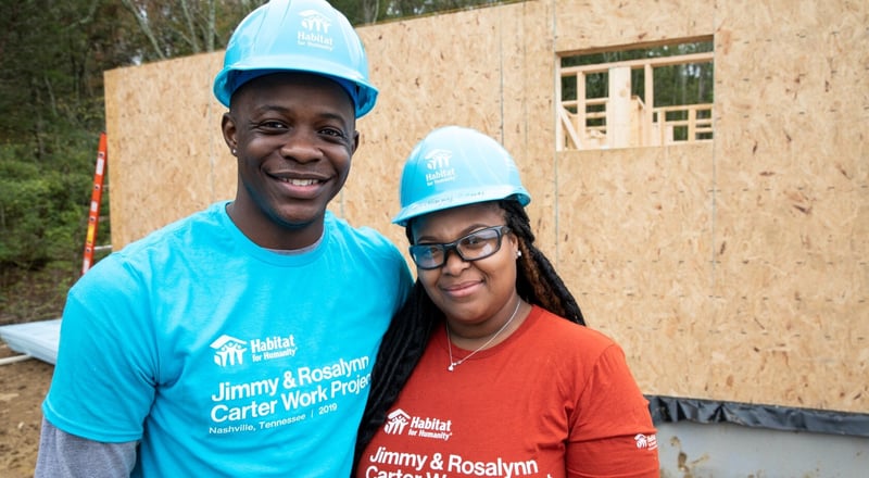 A volunteer and a homeowner wearing hard hats and branded t-shirts in front of an under-construction home at the Carter Work Project.