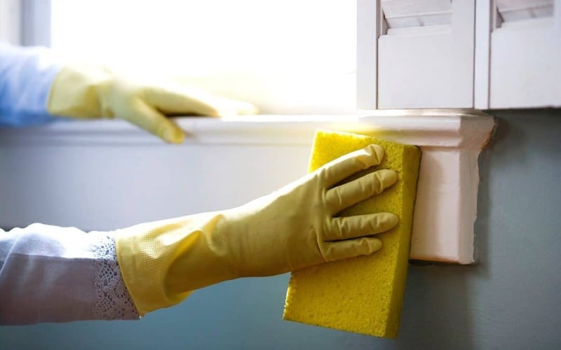 A person wearing yellow cleaning gloves wiping a sponge on a window sill.