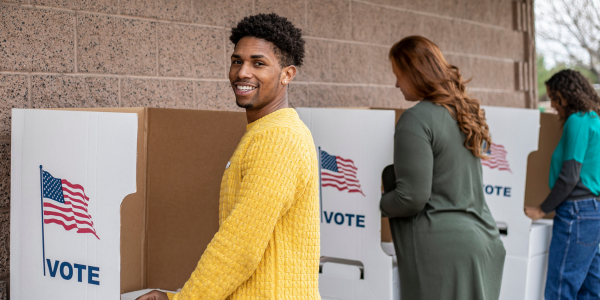 A person in a yellow sweater at a voting booth, smiling at the camera. Two people stand at voting booths in the background.